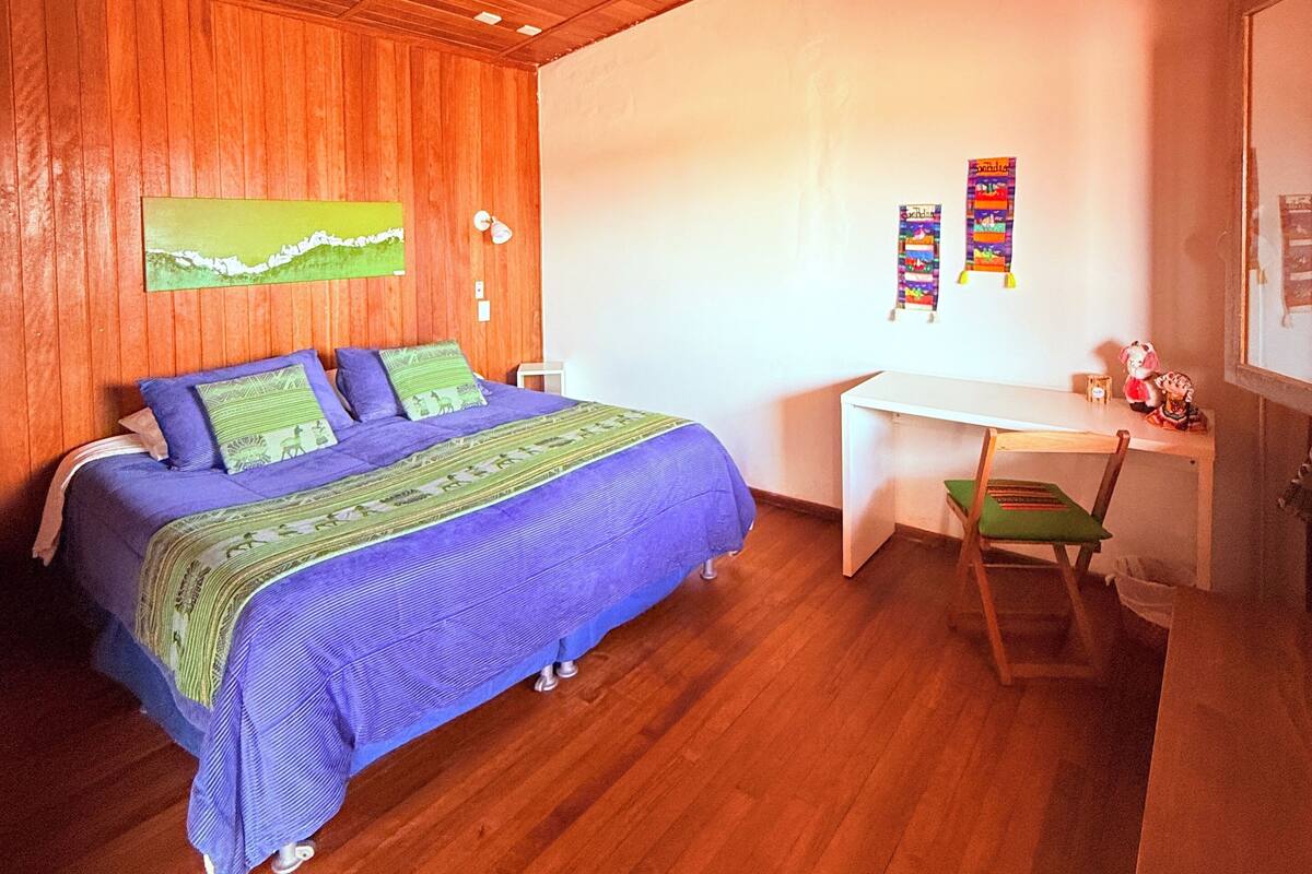 Ein Schlafzimmer mit Holzbalkendecke, einem Bett mit blauer und grüner Bettwäsche, einem Schreibtisch mit Stuhl sowie einer Wand mit farbenfrohen Verzierungen.