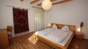 Une chambre avec un lit en bois, une table de chevet avec une lampe, une chaise, une tapisserie murale et une grande fenêtre avec des rideaux.