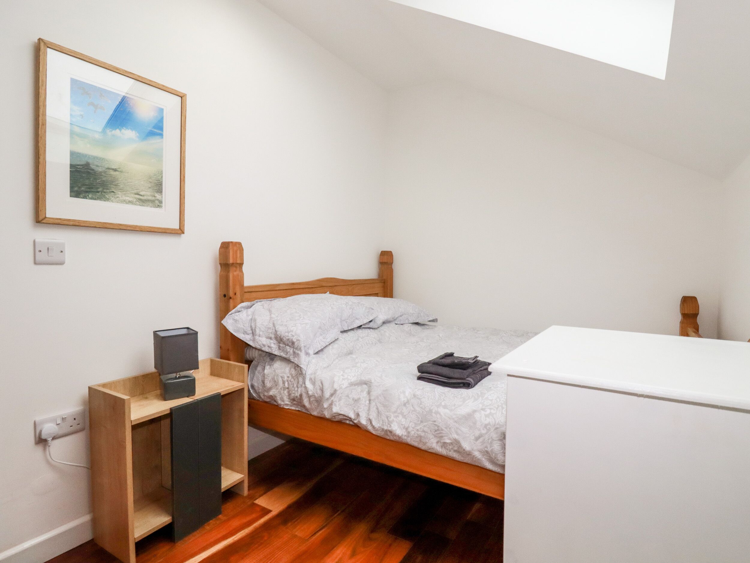 A small, neatly organized bedroom with a wooden bed, a bedside table with a lamp, a framed picture on the wall, and a small white desk.