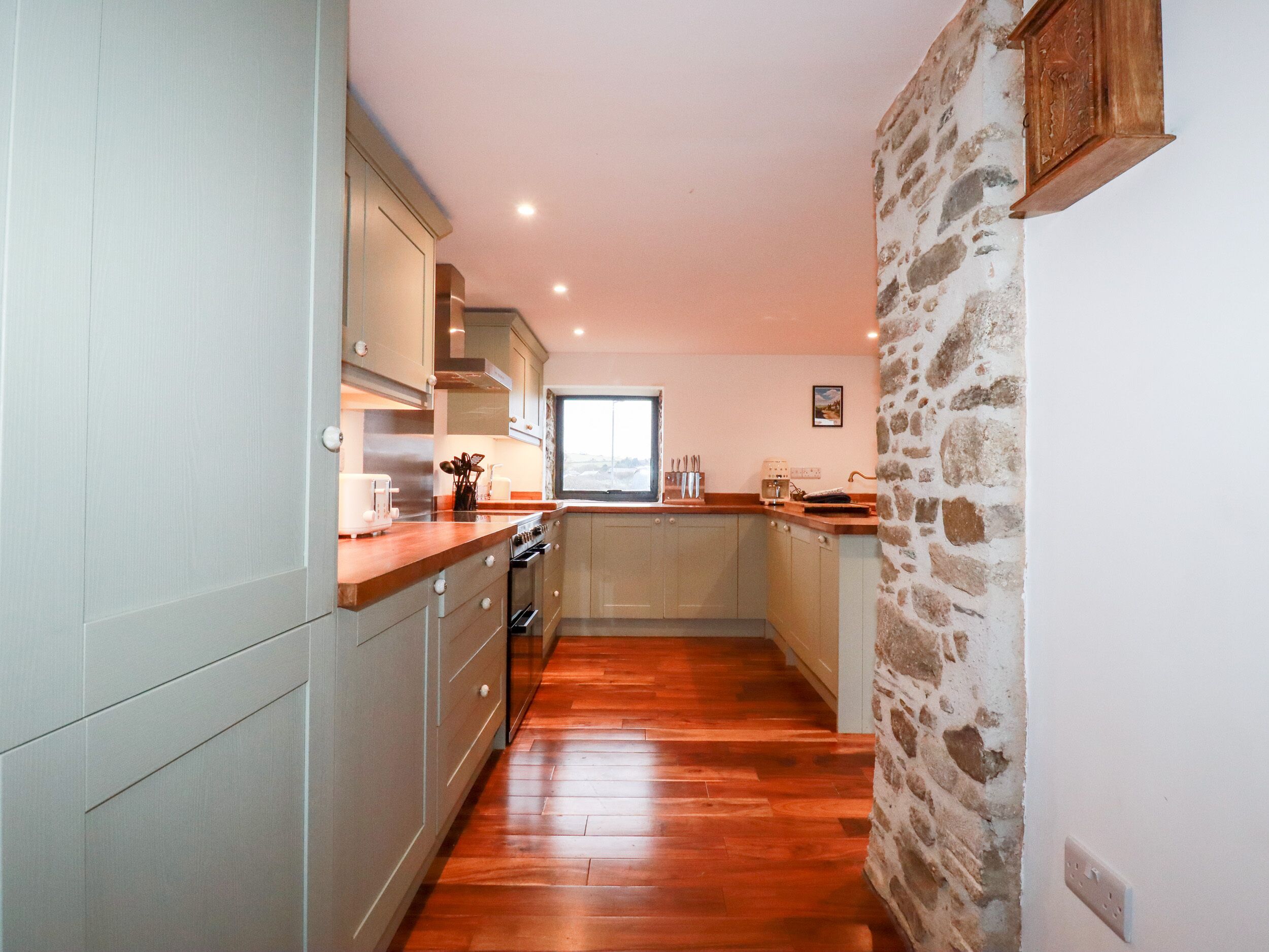 Una cocina estrecha con suelo de madera, armarios de color claro, pared de piedra y una ventana que deja entrar luz natural.
