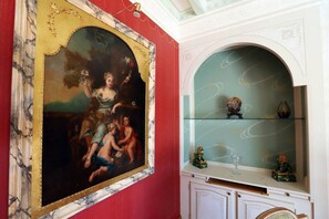 A room with a classical painting on the wall, a glass display cabinet with decorative items, and a red accent wall.