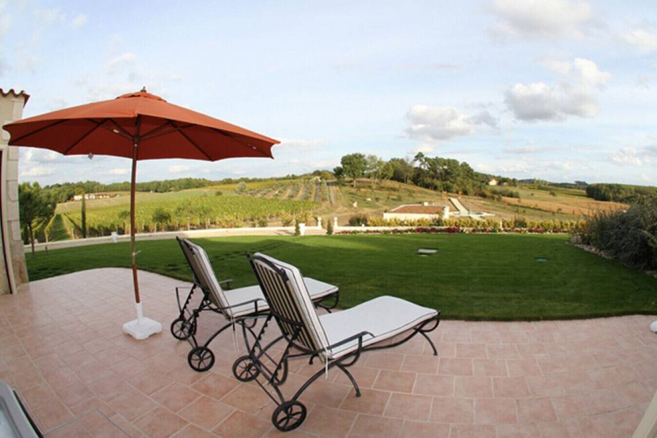 A patio with two chairs and a red umbrella, overlooking a landscaped garden and vineyard.