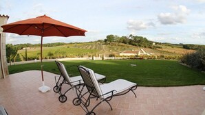 A patio with two chairs and a red umbrella, overlooking a landscaped garden and vineyard.