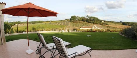 A patio with two chairs and a red umbrella, overlooking a landscaped garden and vineyard.
