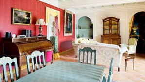 A room with a red wall, a wooden desk, a table, and chairs. There is a chandelier, a cabinet, and a painting on the wall.