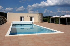A rectangular swimming pool with a tiled deck, a stone wall on one side, and a building with two doors and a small window in the background.