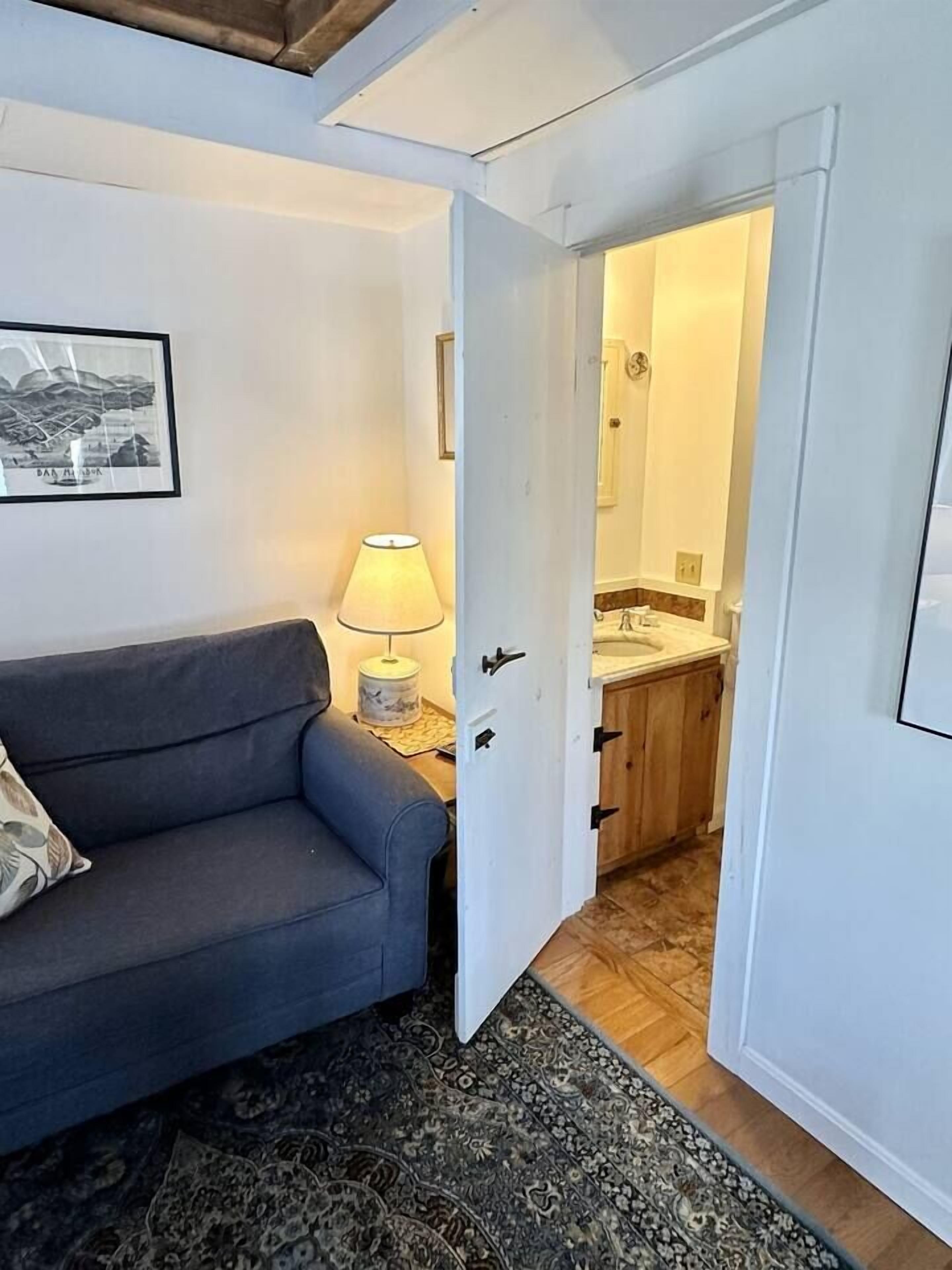 Een kamer met een bank, een lamp, een kamerbreed tapijt en een deur naar een badkamer met een wastafel en spiegel.