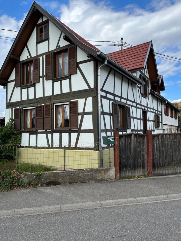 Ein zweistöckiges, traditionelles Fachwerkhhaus mit rotem Ziegeldach, umzäunter Garten und Blick auf die Straße.