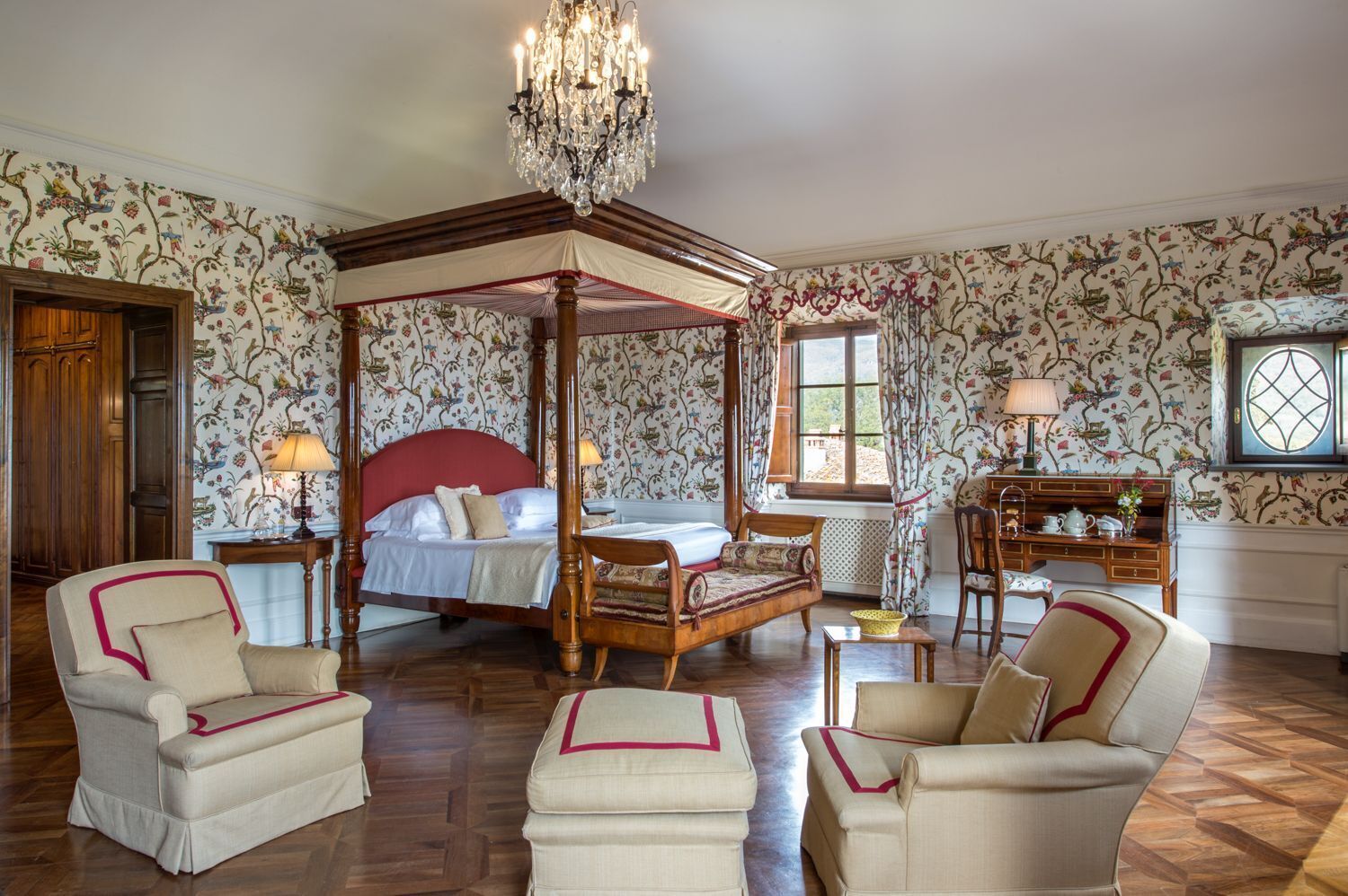 A four-poster canopy bed with a red headboard, a chandelier, and floral wallpaper. There are two armchairs and a small table with a lamp.