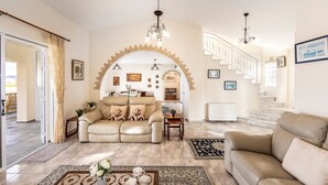 A spacious living room with a beige sofa, a coffee table with decorative items, a patterned rug, and a staircase with a railing leading to an upper level. - Villa Chrysos - Three Bedroom Villa, Sleeps 6 (Tala)