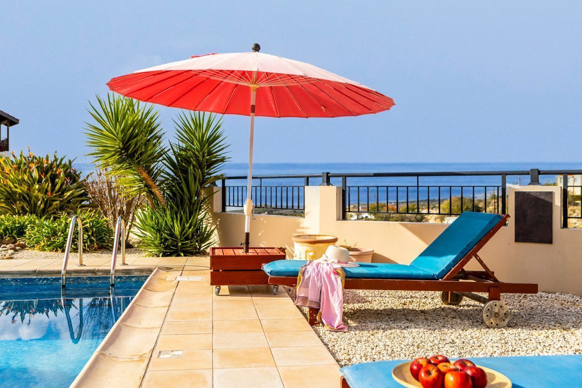 A poolside area with a red umbrella, a lounge chair, and a table with fruit.