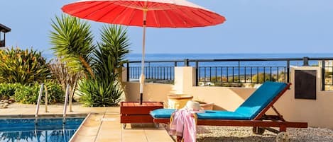 A poolside area with a red umbrella, a lounge chair, and a table with fruit.