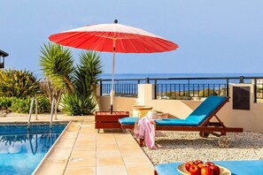 A poolside area with a red umbrella, a lounge chair, and a table with fruit. - Villa Chrysos - Three Bedroom Villa, Sleeps 6 (Tala)