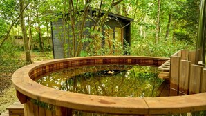 Una bañera de hidromasaje de madera con una terraza de madera alrededor, ubicada en un área boscosa con un cobertizo pequeño al fondo.