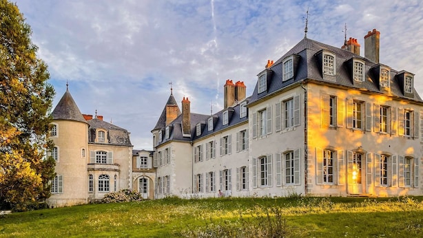 Un edificio grande e histórico con múltiples torres y chimeneas, rodeado por un césped cuidado y árboles.