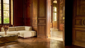 Una habitación con piso de madera en patrón de espiga, un sofá seccional color beige y una mesa de centro de vidrio con flores secas.