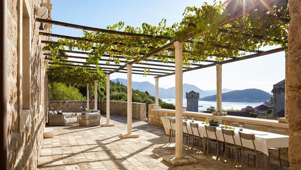 An outdoor patio with stone flooring, a pergola with vines, and a table set for dining with a scenic view of mountains and water. - Villa Reverie - Seven Bedroom Villa, Sleeps 14 (Sudurad)