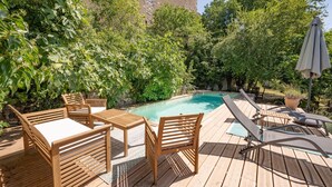 Een houten terras met een zwembad, omgeven door weelderige begroeiing en een landelijk gebouw op de achtergrond.