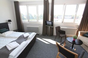 A hotel room with a bed, a desk, a chair, a small table with a plant, and a TV on the wall.