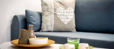 Una sala moderna con un sofá azul, un cojín decorativo con una cita, una mesita redonda con una bandeja que sostiene un florero, un cuenco y un vaso de bebida verde.
