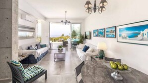 A modern living room with a sofa, a coffee table, and a dining table. There are framed pictures on the wall and a view of the outdoors. - Latchi Infinity Blue (Neo Chorio)