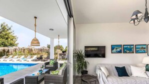 A modern living room with a pool, wicker furniture, a sofa, and a TV wall-mounted above a sofa. - Latchi Infinity Blue (Neo Chorio)
