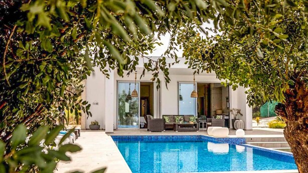 A modern pool area with a clear blue pool, surrounded by a white building with large windows and a seating area with outdoor furniture. - Latchi Infinity Blue (Neo Chorio)