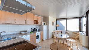 A compact kitchen with a built-in microwave, a coffee maker, and a washing machine. There is a dining area with a table and chairs, and a large window providing natural light.