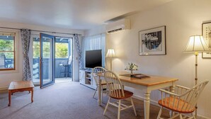 A room with a dining table, chairs, a television, and a balcony with curtains. - 1BR Waterfront Condo in Downtown BH [Ocean Breeze] (Bar Harbor)