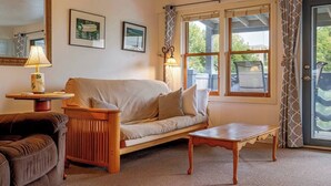 A living room with a sofa, a coffee table, a lamp, and a window with curtains. - 1BR Waterfront Condo in Downtown BH [Ocean Breeze] (Bar Harbor)