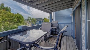 A covered outdoor patio with a table and chairs, a blue building in the background, and a view of trees and hills. - 1BR Waterfront Condo in Downtown BH [Ocean Breeze] (Bar Harbor)