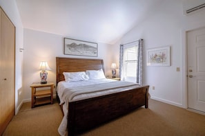 Quarto com uma cama de madeira, duas mesas de cabeceira com abajures, uma janela com cortinas e uma porta.