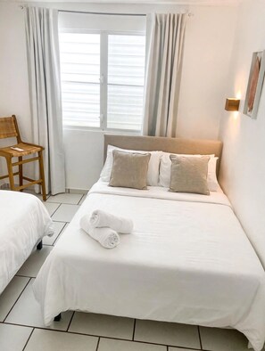 Ein Hotelzimmer mit einem Bett, zwei Kissen, einem Sessel, einem Fenster mit Vorhängen und einer Wandleuchte.