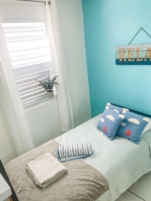 Une chambre à coucher avec un lit, des oreillers, une serviette, une fenêtre avec des stores et un panneau décoratif sur le mur.