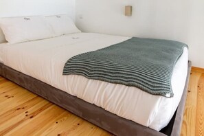 Un lit avec un matelas blanc et une couverture grise, un plancher en bois et un mur blanc agrémenté d’un petit luminaire.