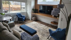 A modern living room with a large window, a flat-screen TV, a ceiling fan, and a dining area with a table and chairs. -  NEW Build-777 Hideaway (Grants Pass)