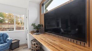 A modern room with a large black TV on a wooden wall, a blue chair, a small table with a plant, and a basket. -  NEW Build-777 Hideaway (Grants Pass)