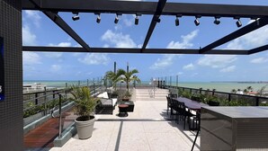 An outdoor terrace with a view of the ocean, featuring seating areas, potted plants, and a railing. - Flat Premium Ozean Home & Service - João Pessoa (João Pessoa)