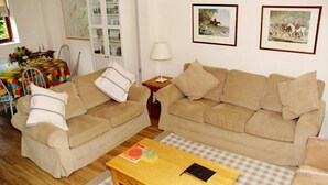 Un salon avec deux canapés beige, une table basse en bois et un tapis à carreaux. Des tableaux encadrés ornent le mur, et on aperçoit, au fond, une vitrine.