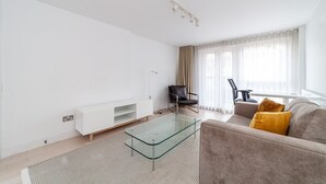 A modern living room with a glass coffee table, a sofa with cushions, a chair, and a TV stand.