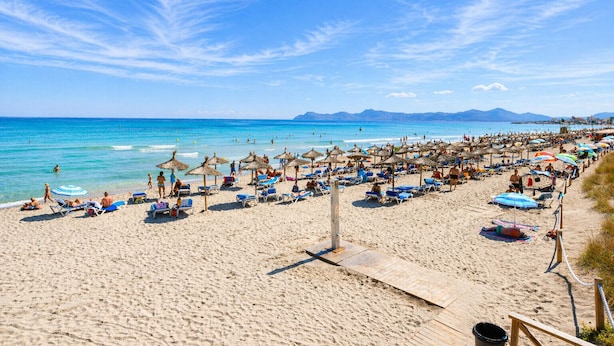A beach with numerous sun loungers and umbrellas, a wooden walkway, and a clear view of the sea and distant mountains.