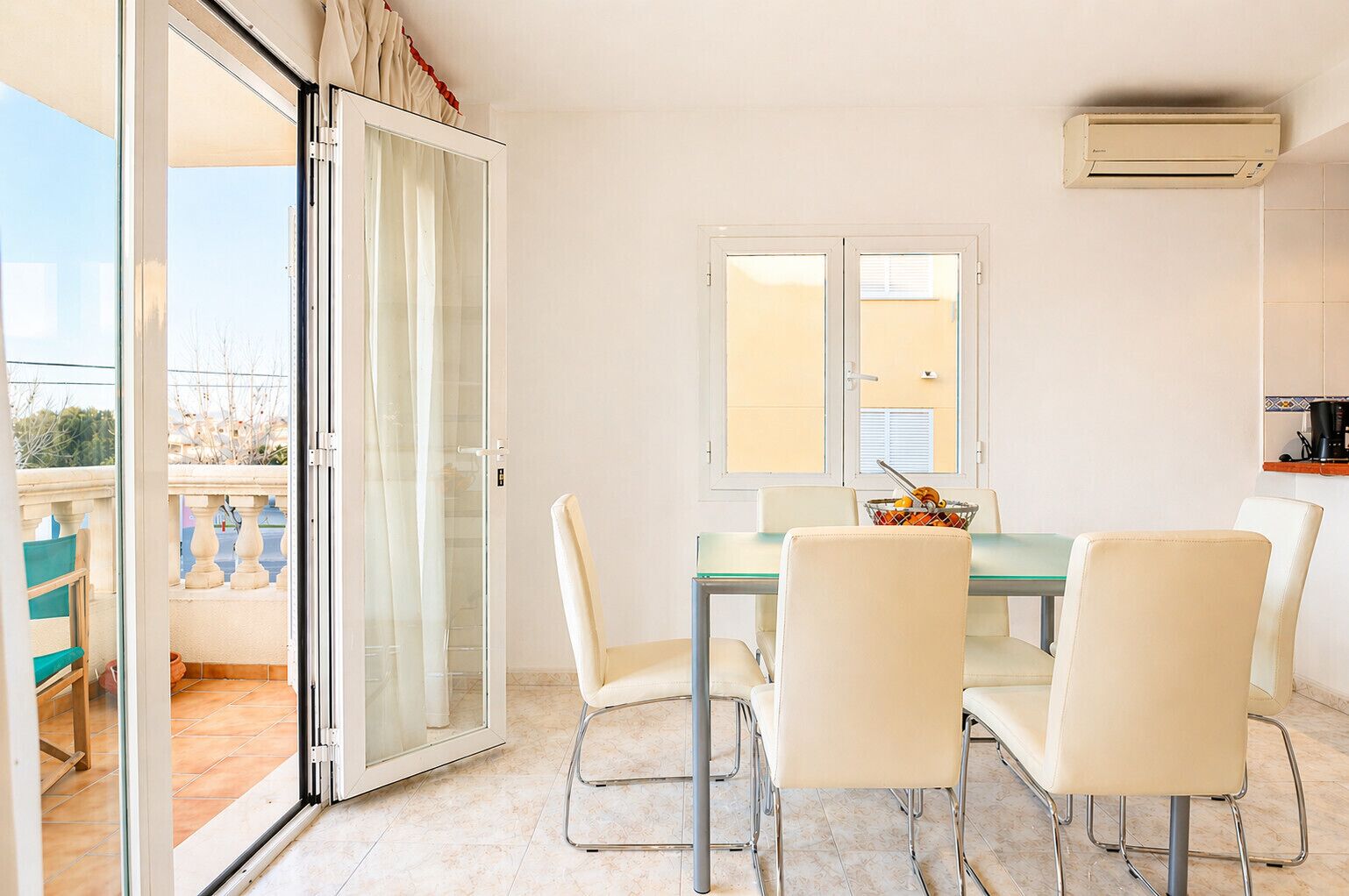 A dining area with a glass table and four chairs, a window with blinds, and an air conditioning unit on the wall.