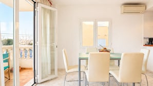 A dining area with a glass table and four chairs, a window with blinds, and an air conditioning unit on the wall.