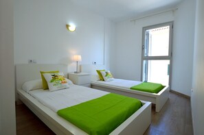 A hotel room with two beds, a nightstand, a lamp, and a window with blinds. - Apartamento en Sa Marina cerca de la playa (Alcúdia)
