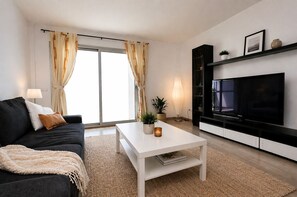 A modern living room with a sofa, coffee table, TV, and a large window with curtains. - Apartamento en Sa Marina cerca de la playa (Alcúdia)
