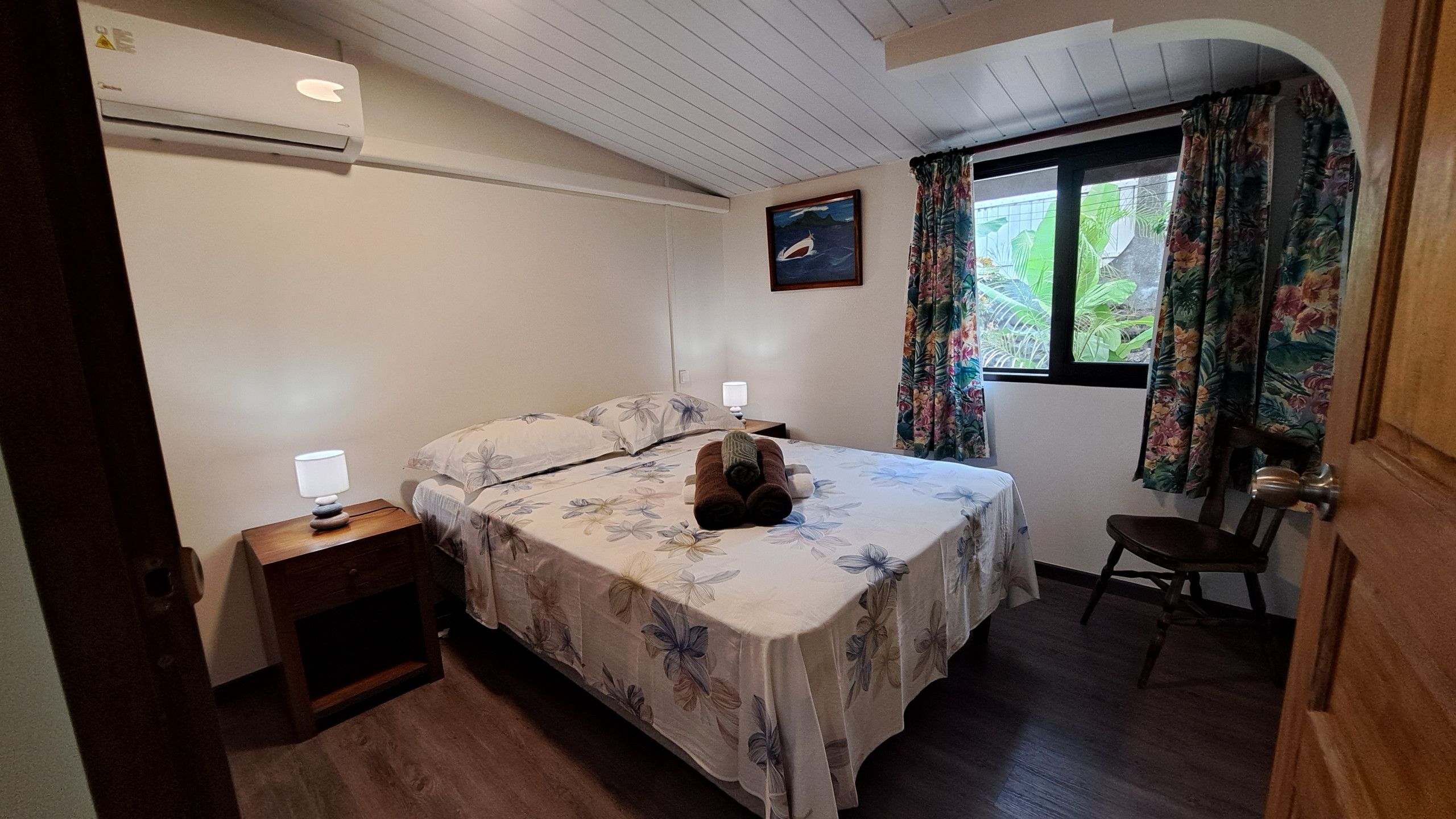 A bedroom with a bed, a nightstand, a chair, a window with curtains, and a painting on the wall.