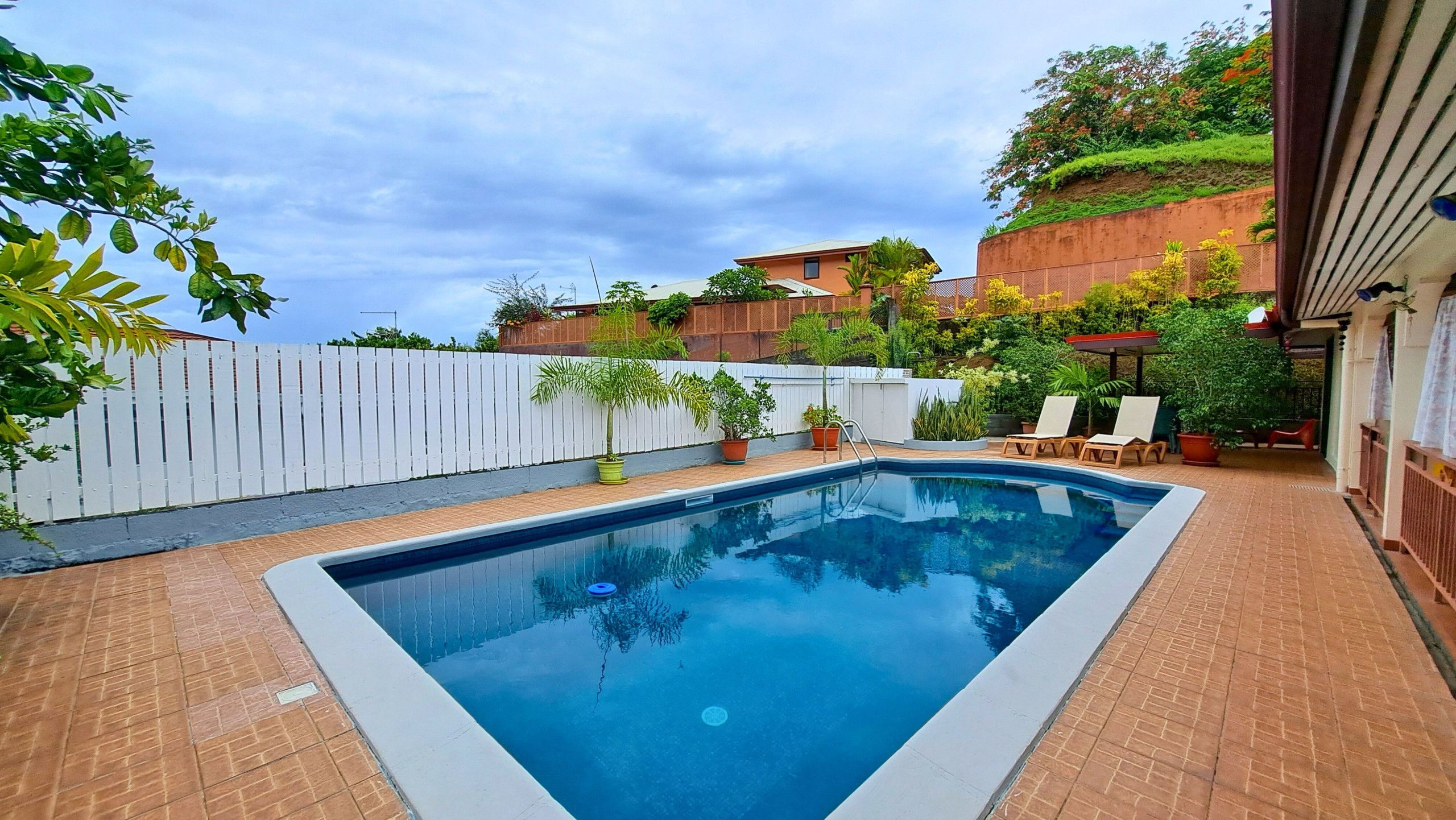 A rectangular swimming pool with a white border, surrounded by a brick patio. There are potted plants and a white fence in the background.