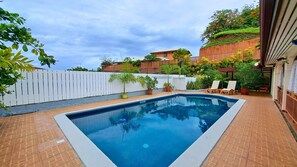 A rectangular swimming pool with a white edge, surrounded by a brick patio. There are lounge chairs and potted plants around the pool.