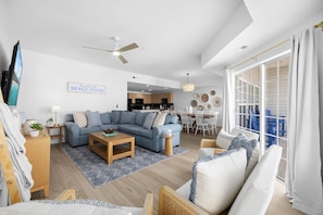 A spacious living room with a large sectional sofa, a wooden coffee table, and a dining area with a white table and chairs. A ceiling fan and a sign reading "Beach House" are visible.