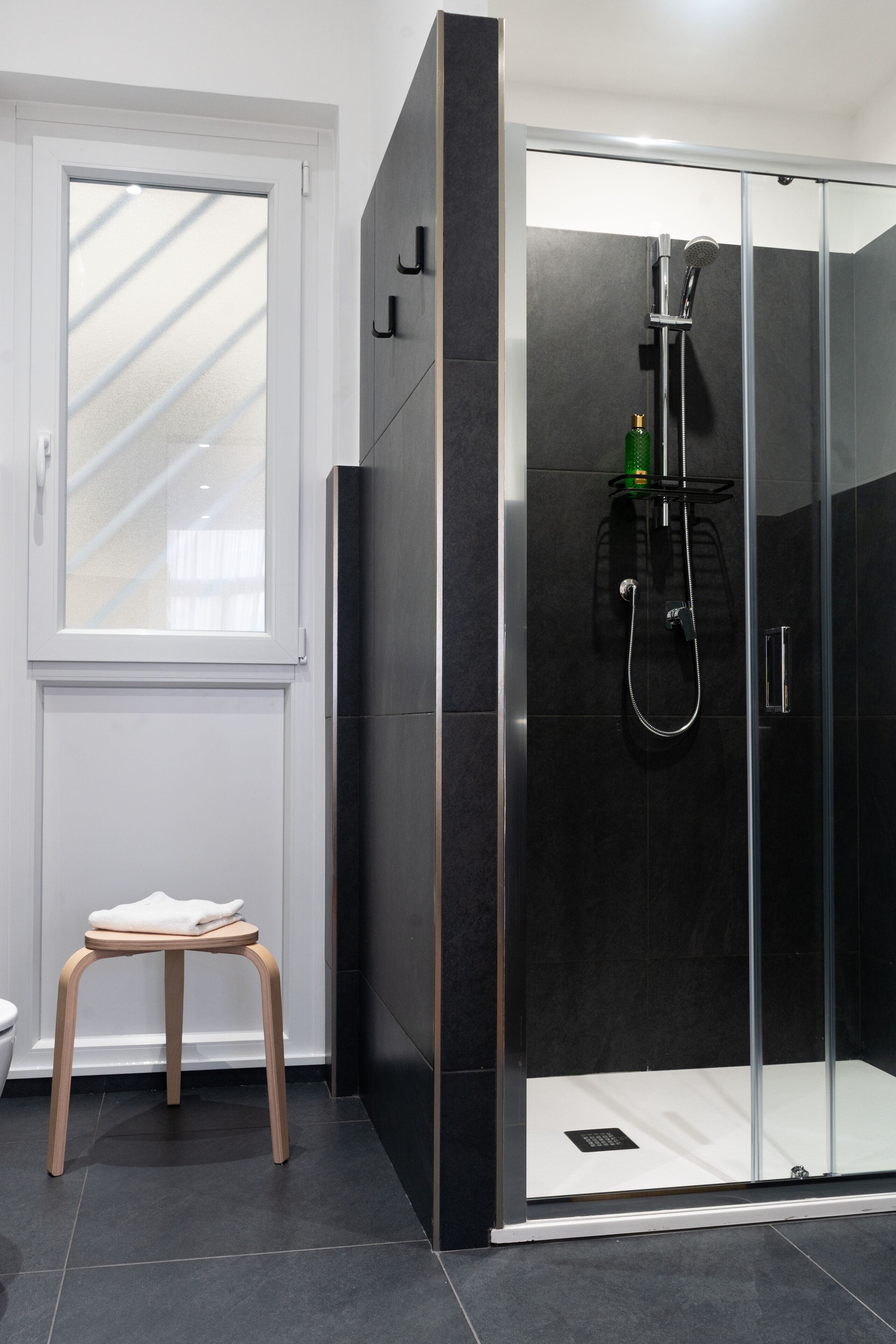 Een moderne badkamer met een douchekabinet van glas, een houten kruk en een raam dat voor natuurlijk licht zorgt.